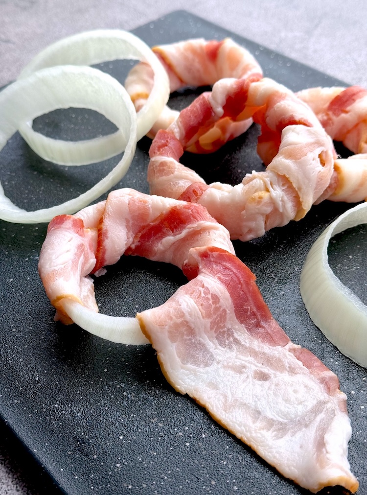 Bacon wrapped around onion rings ready to go into the oven or air fryer for a tasty snack. Once baked they're crispy and delicious. Brush with your favorite bbq sauce to finish them off. Crunchy, savory, sweet, and irresistible!