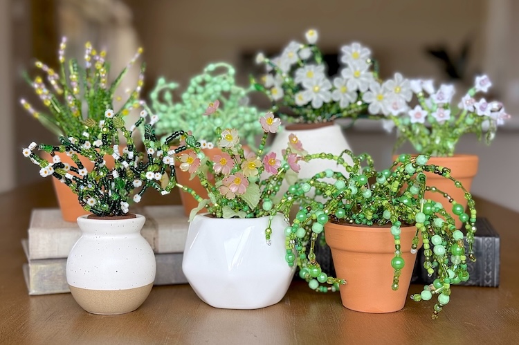 Variety of DIY beaded plants sitting together on a table made with jewelry beads, foam, clay pots, and wire. 
